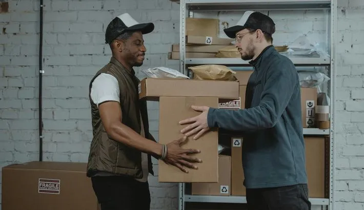 Two warehouse workers handing each other a large cardboard box against the backdrop of racks of parcels